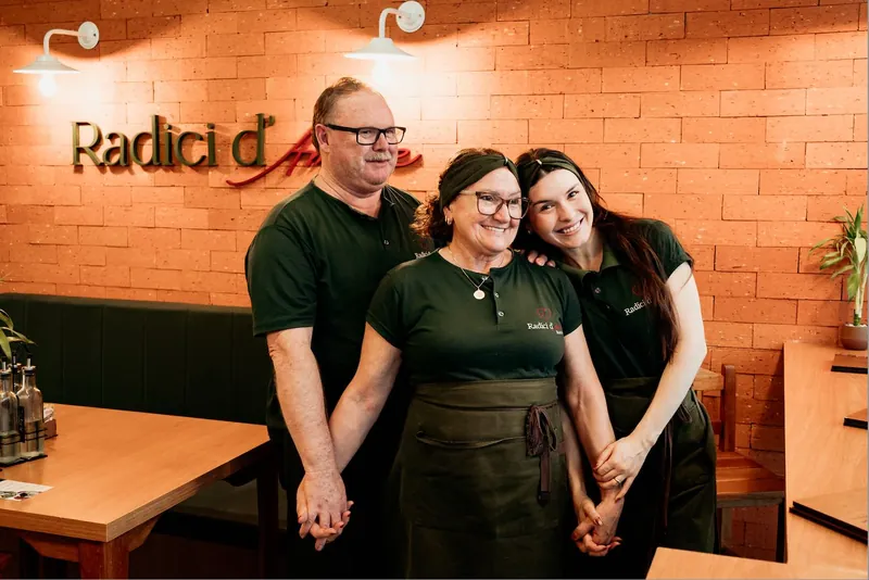 A família Radici d'Amore sorrindo, de uniforme verde, diante da parede de tijolos com o letreiro do bistrô